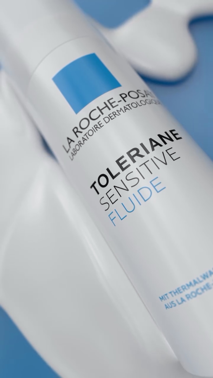 La Roche-Posay Toleriane Sensitive Fluide bottle at an angle, surrounded by flowing white cream on a blue surface