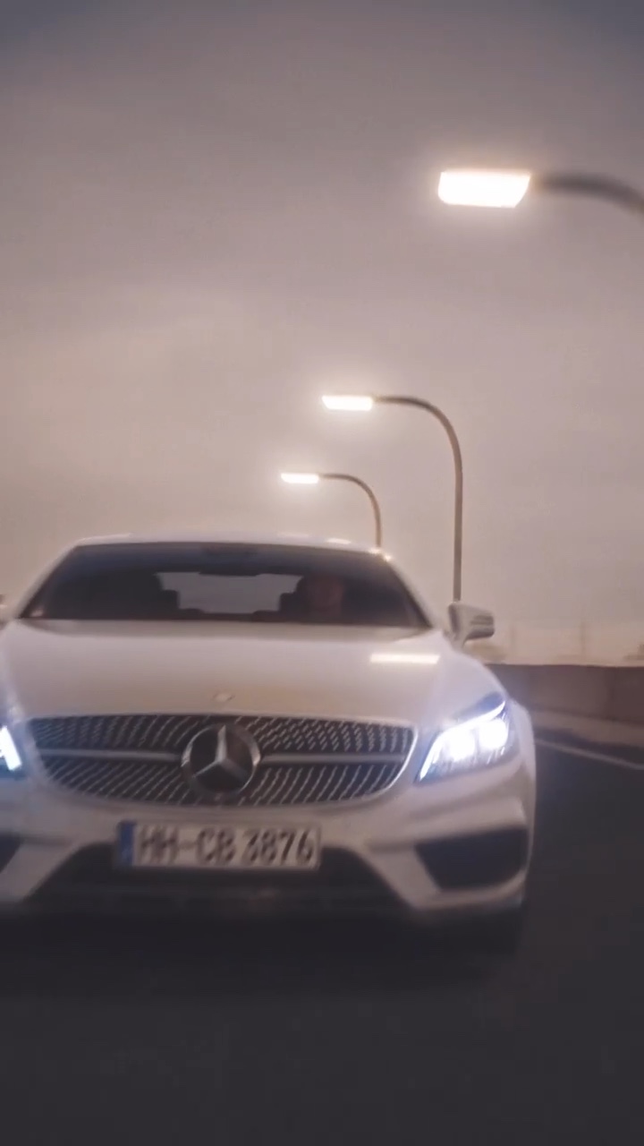 Frontansicht eines silbernen Mercedes CLS mit eingeschalteten Scheinwerfern auf einer Straße in der Dämmerung, im Hintergrund Straßenlaternen